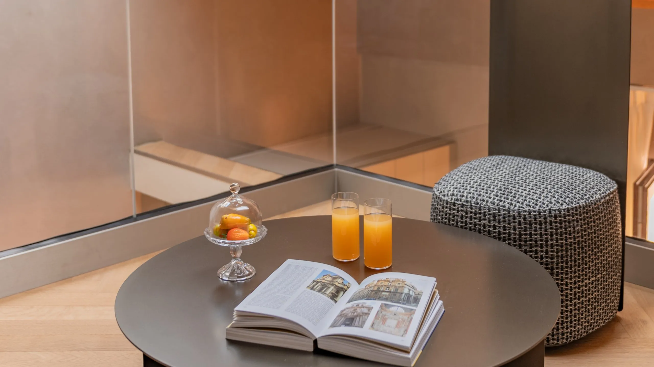 Angolo lettura Suite Sitwell: tavolino tondo, succhi freschi, libro e pouf su soppalco con vetrata a Villa Conrad.