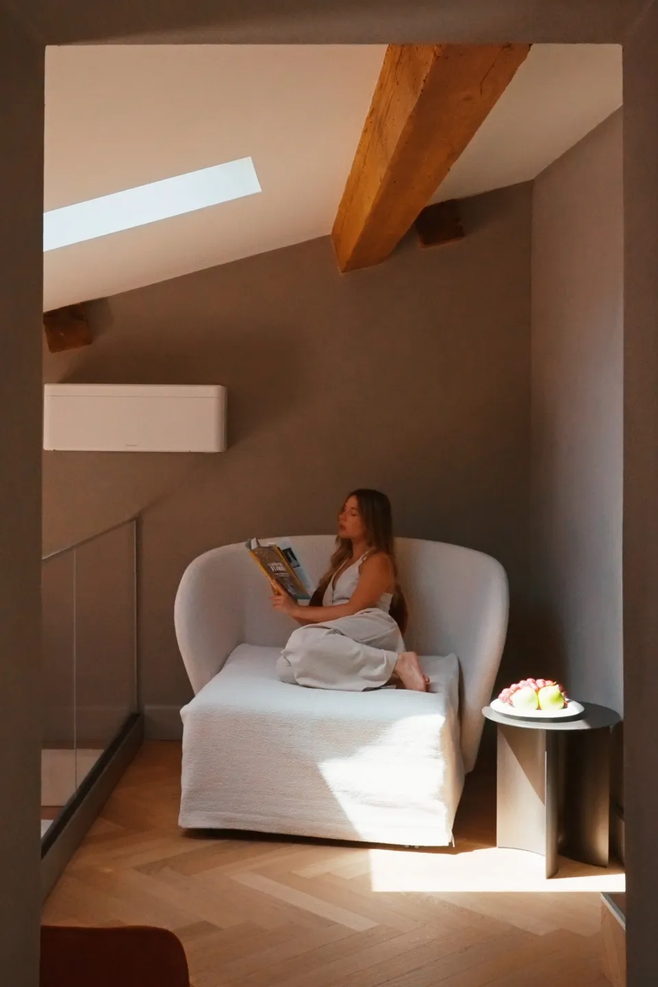 Relax al soppalco di Villa Conrad Taormina: donna legge su poltrona di design sotto travi in legno e luce zenitale.