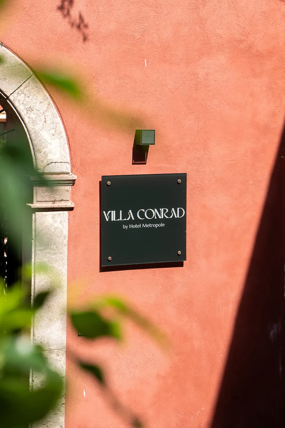 Insegna Villa Conrad by Hotel Metropole a Taormina: dettaglio logo su parete rosso pompeiano e arco in pietra.